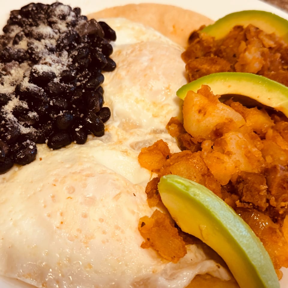Close-up of a diner breakfast with fried eggs, black beans, seasoned potatoes, and avocado slices.