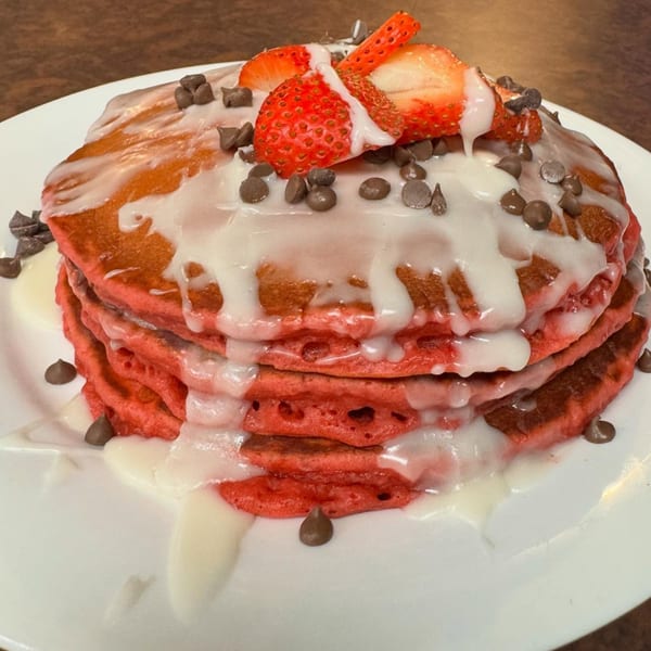 Stack of red velvet pancakes with white icing, chocolate chips, and sliced strawberries on a white plate.