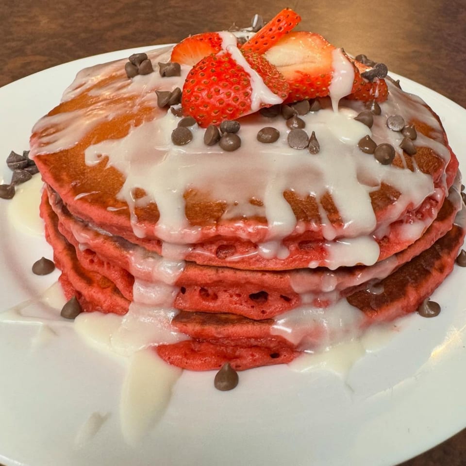 Stack of red velvet pancakes with white icing, chocolate chips, and sliced strawberries on a white plate.