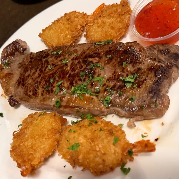 Grilled steak with crispy fried shrimp and a side of sweet chili sauce on a white plate.